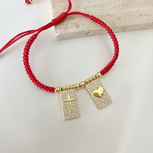 Adjustable red cord bracelet with crystal-studded charms, including a gold cross and heart, displayed on a neutral stone surface.