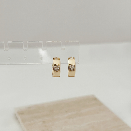 A pair of gold huggie hoop earrings with a small heart-shaped crystal design in the center, hanging from a clear acrylic display stand.
