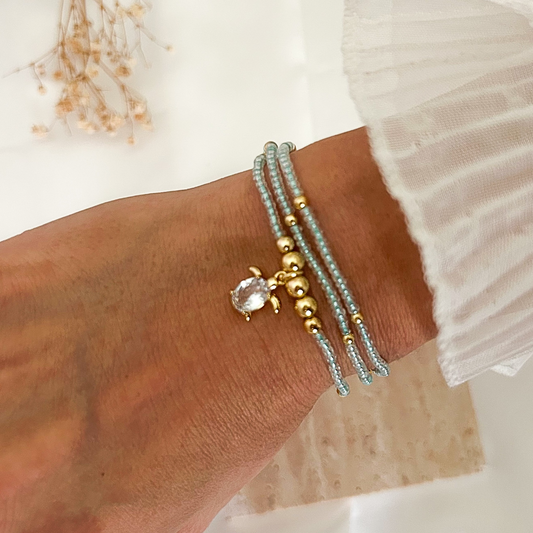 Handmade turquoise beaded bracelet with gold details and a crystal turtle charm, worn on the wrist and styled with a white ruffled sleeve against a soft neutral background.