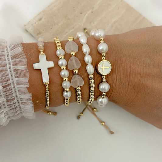 Close-up of layered gold bracelets with pearls, pink heart beads, and meaningful cross charms.
