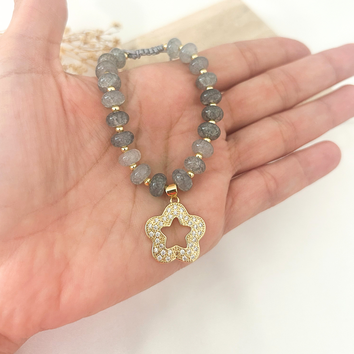 A beaded stretch bracelet with gray stone beads and gold-toned accents, featuring a gold four-petal flower pendant paved with small clear stones, shown resting on an open hand.