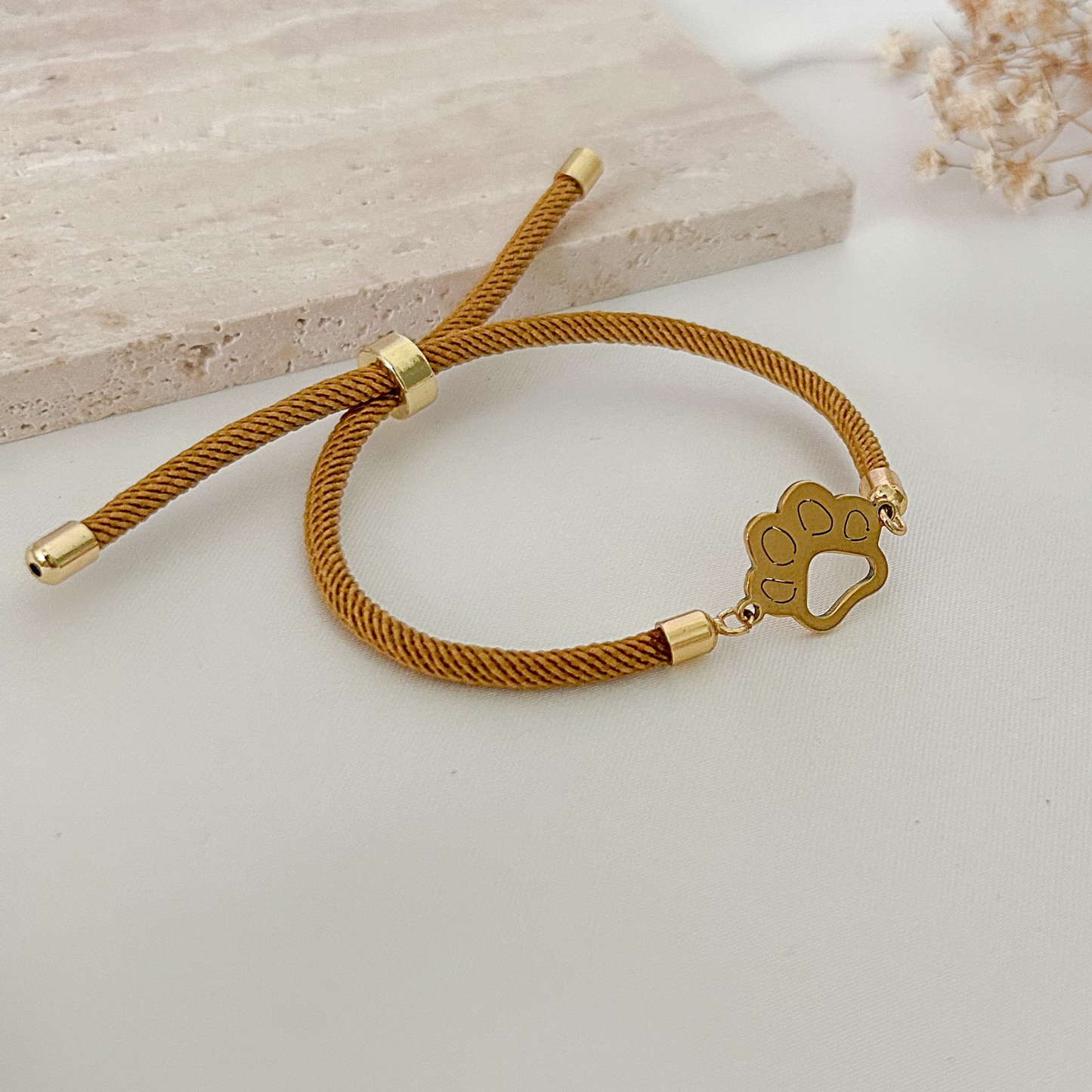 Minimalist pet lover bracelet featuring a gold-tone paw charm on a camel-colored adjustable cord, styled on a neutral stone surface with soft dried floral accents.