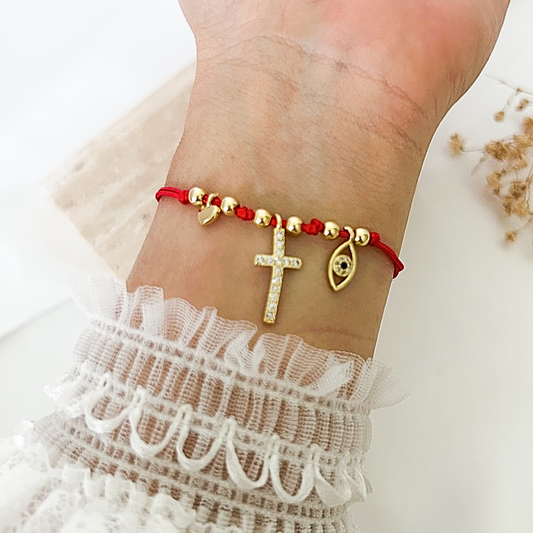 Close-up of a red protection bracelet worn on the wrist, accented with a sparkling gold cross, evil eye charm, and heart detail, paired with a delicate white lace sleeve.