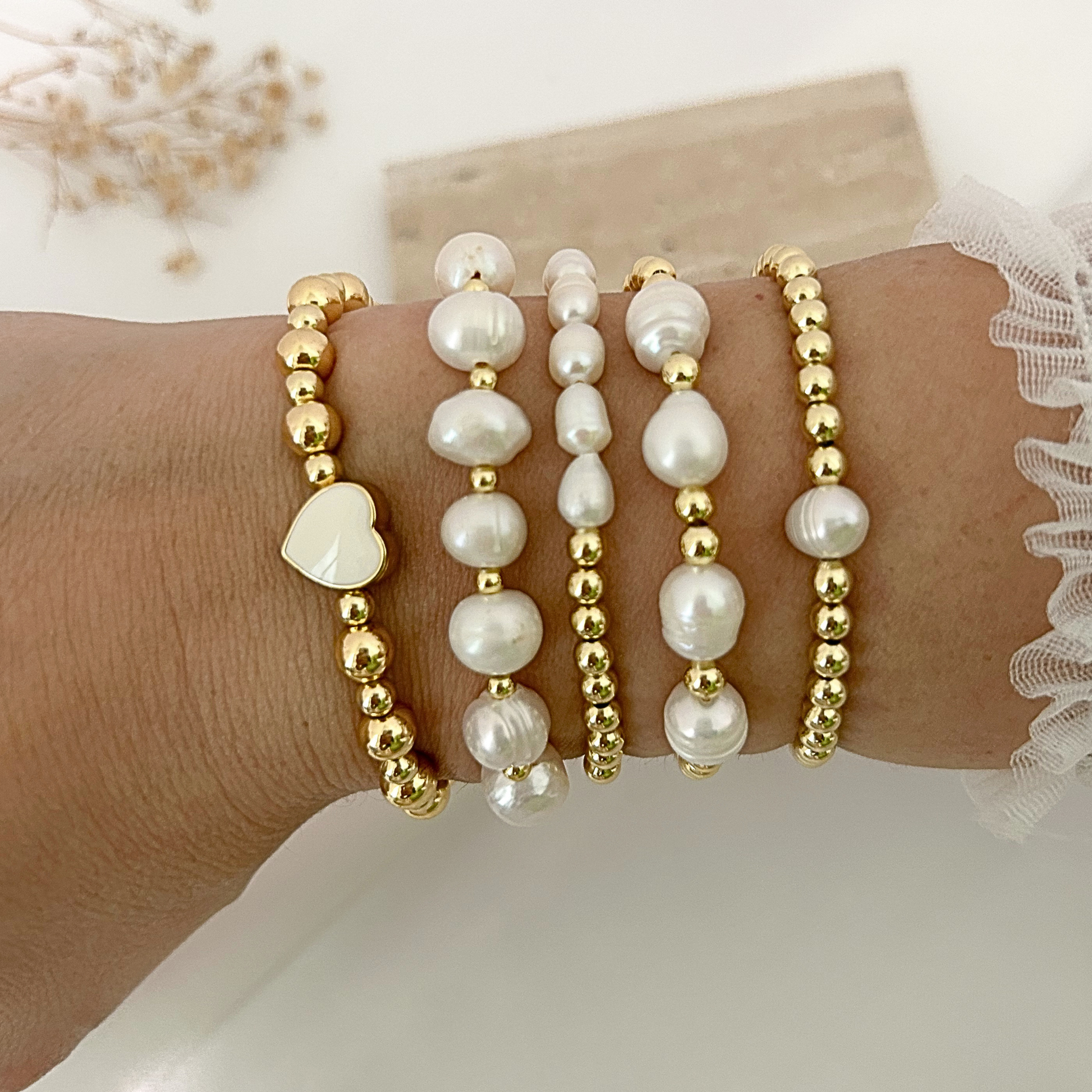 Four beaded and pearl bracelets in gold and white tones, shown stacked on a person's wrist. One bracelet features a white heart-shaped shell charm.