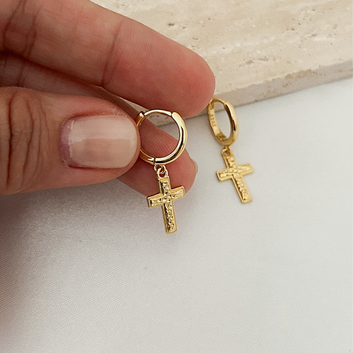A close-up of a hand holding a pair of small gold hoop earrings with dangling textured cross charms, set against a light fabric background and a stone slab.