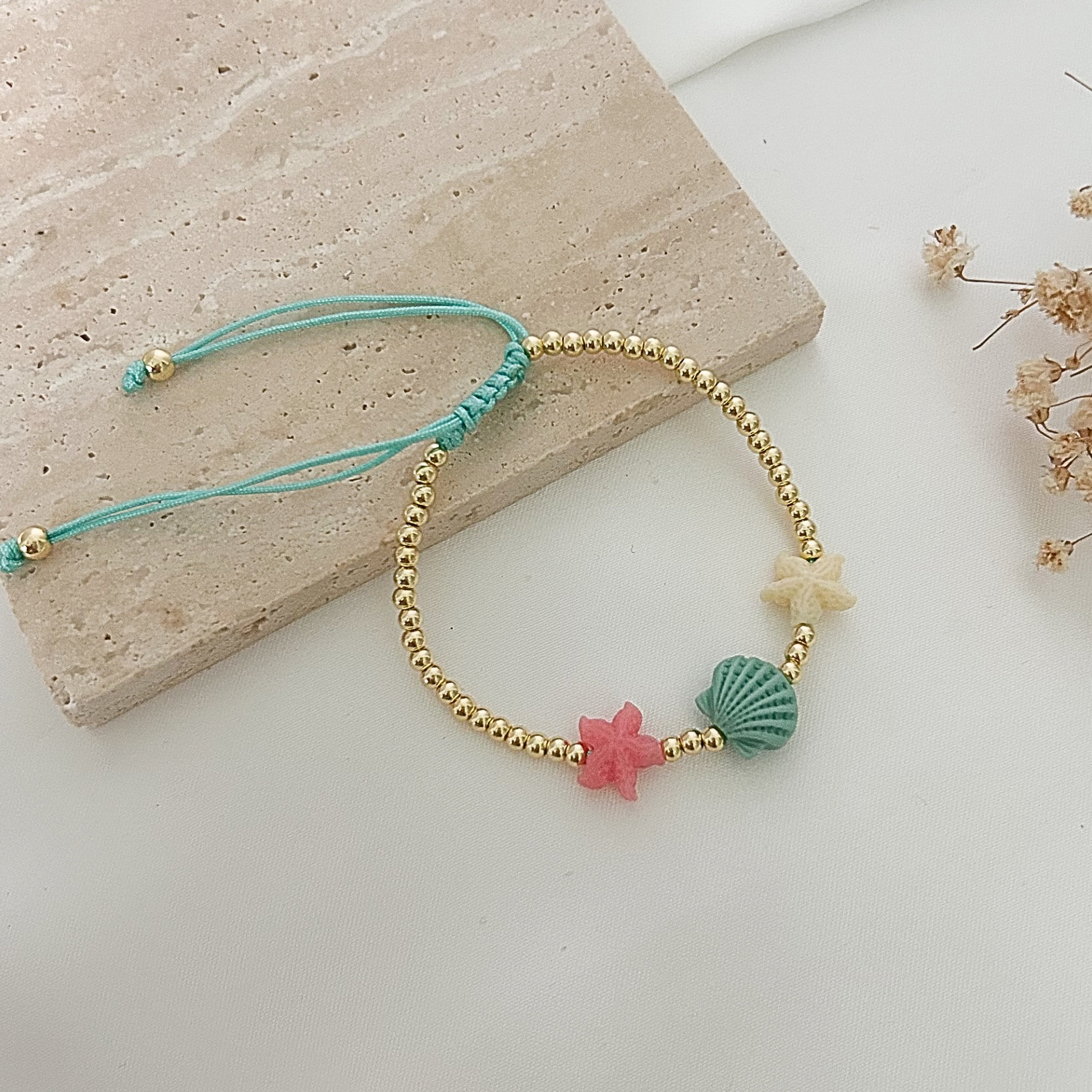 Handmade adjustable bracelet with gold-tone beads and pastel sea-life details, featuring starfish and shell-shaped accents in soft beachy colors, styled on a stone surface with dried flowers.