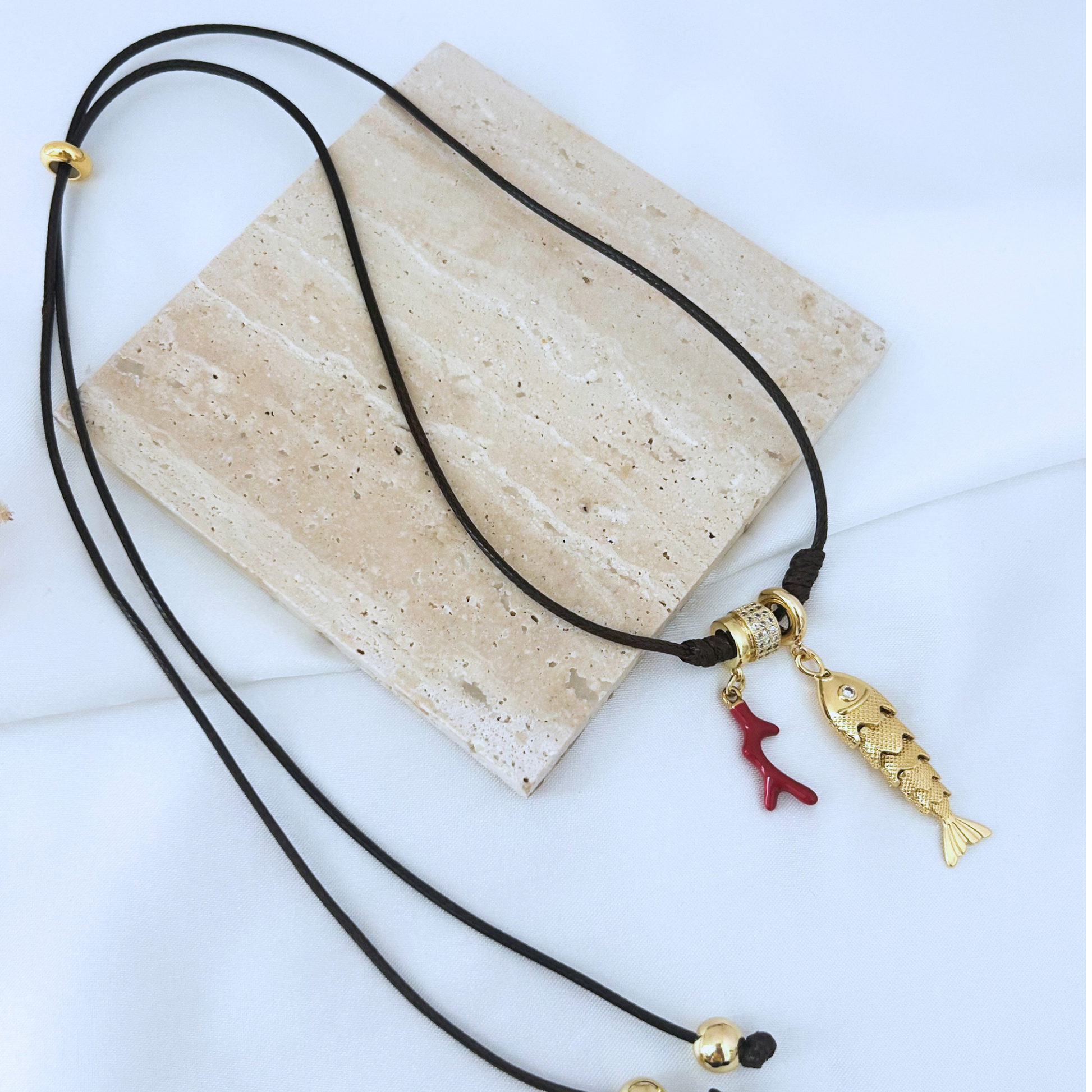 Adjustable nautical-inspired necklace featuring a black cord, sparkling gold-tone slider bead, a detailed gold fish charm, and a vibrant red coral charm. A unique coastal accessory with an elegant Mediterranean style.