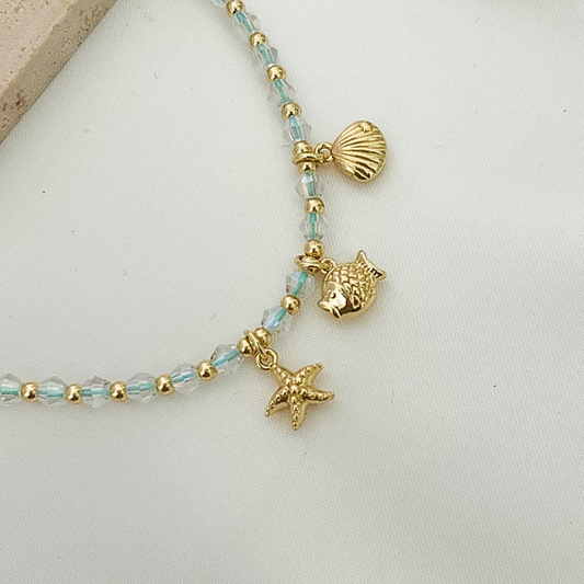 Handmade ocean-themed bracelet featuring faceted crystal beads, gold details, and coastal charms including a starfish, fish, and shell, displayed on a neutral fabric background.