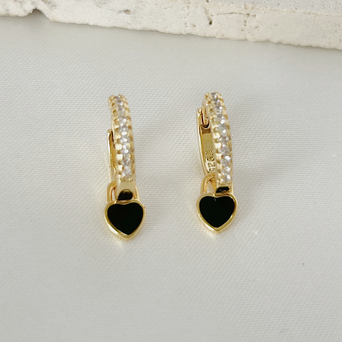 A pair of gold huggie hoop earrings with clear pavé stones and dangling black heart charms, displayed on a textured white surface.