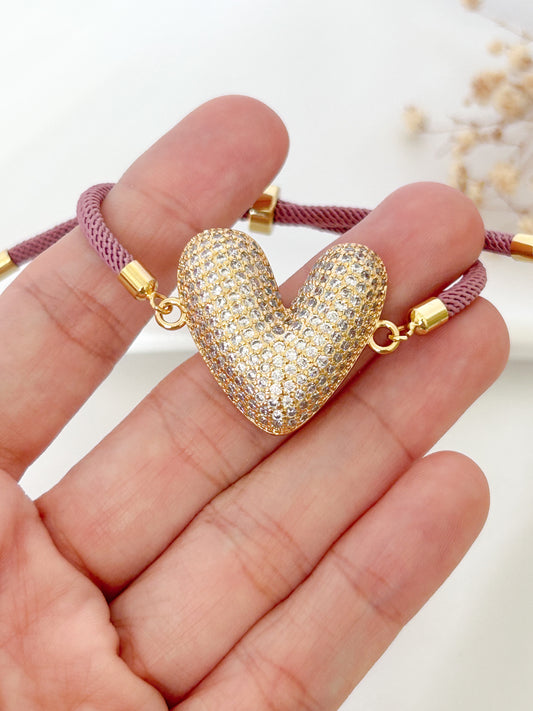 Gold heart-shaped pendant on a pink cord held in a hand against a white background