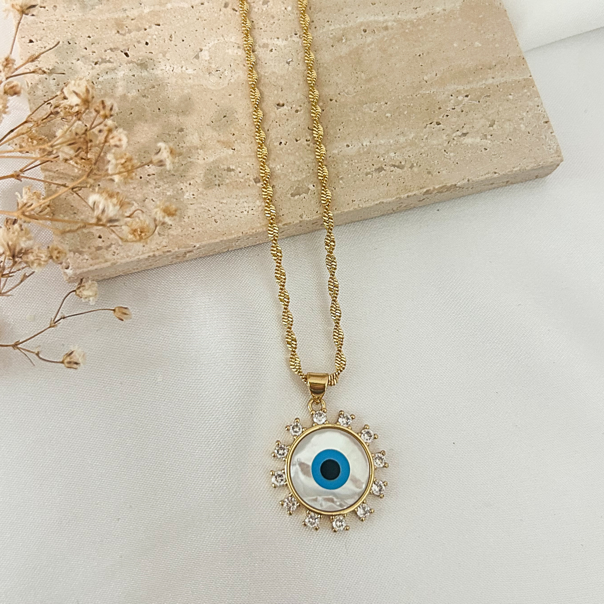 Gold evil eye pendant necklace featuring a mother-of-pearl center, blue enamel eye, sparkling cubic zirconia halo, and textured gold chain, displayed on a neutral minimalist background.