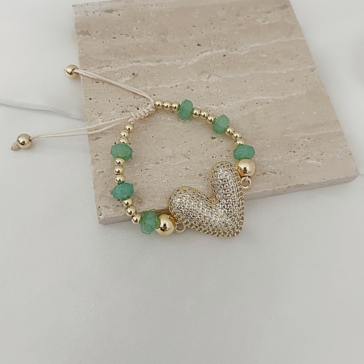 A gold beaded bracelet with green faceted beads and a textured gold heart charm, displayed on a light beige stone tile over a white surface.