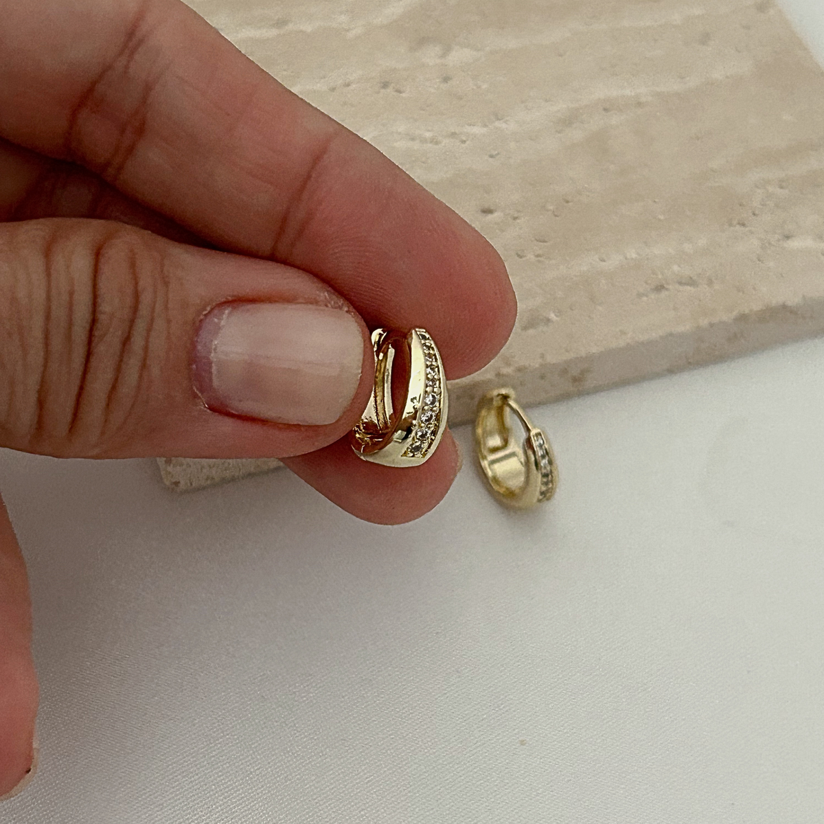 A close-up of a hand holding a small gold huggie hoop earring with tiny clear stones, with a second matching earring resting on a light surface nearby.