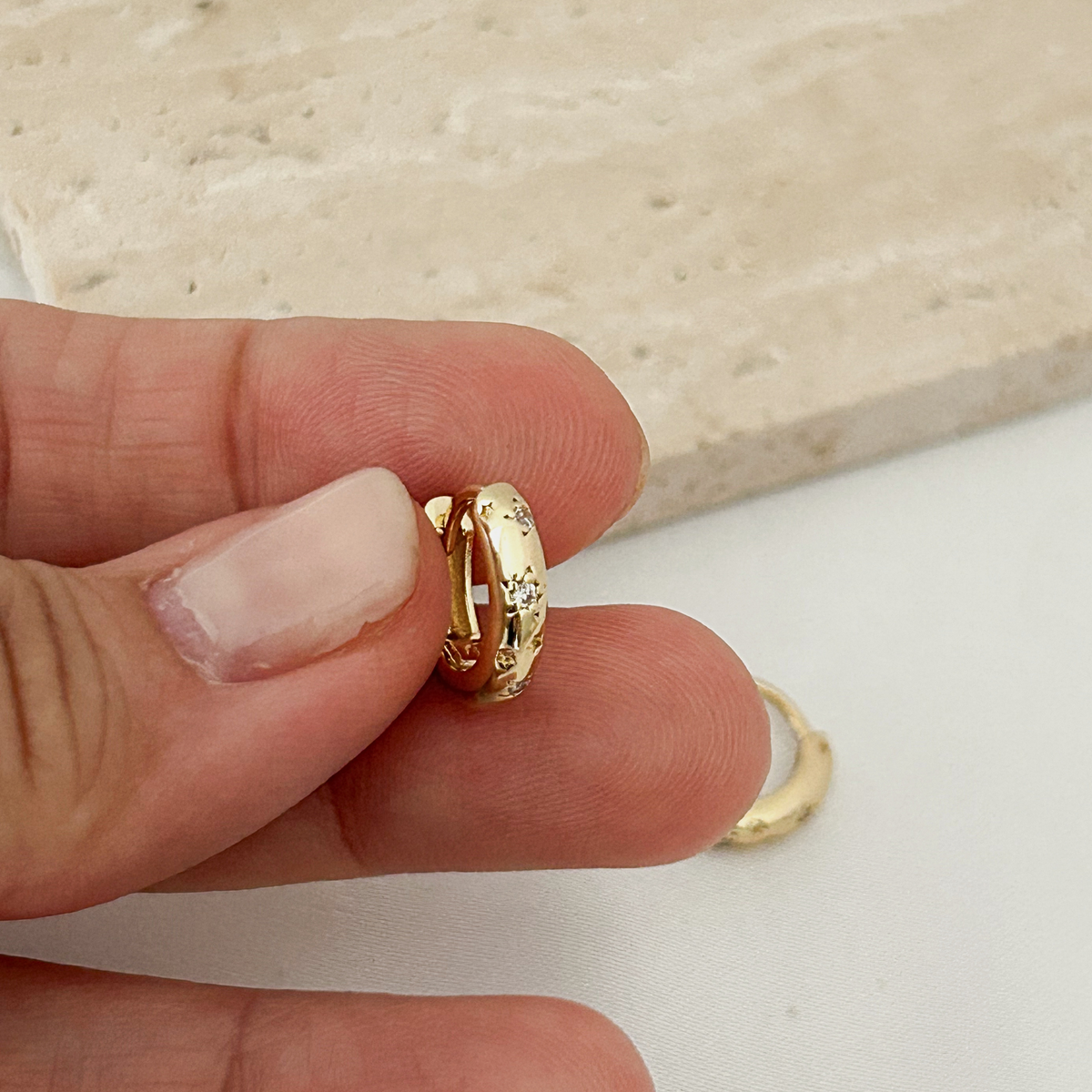 A close-up of a hand holding a small gold huggie hoop earring, with a second matching hoop lying on a white surface in the background.