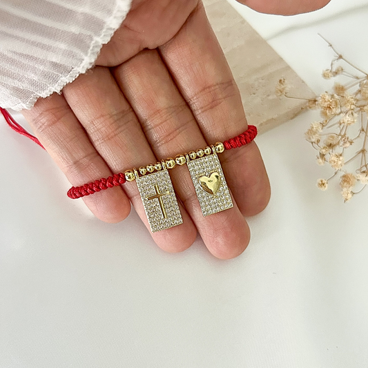 Close-up of a handmade red string bracelet featuring two sparkling rectangular charms with a gold cross and heart design, accented with gold-tone beads.