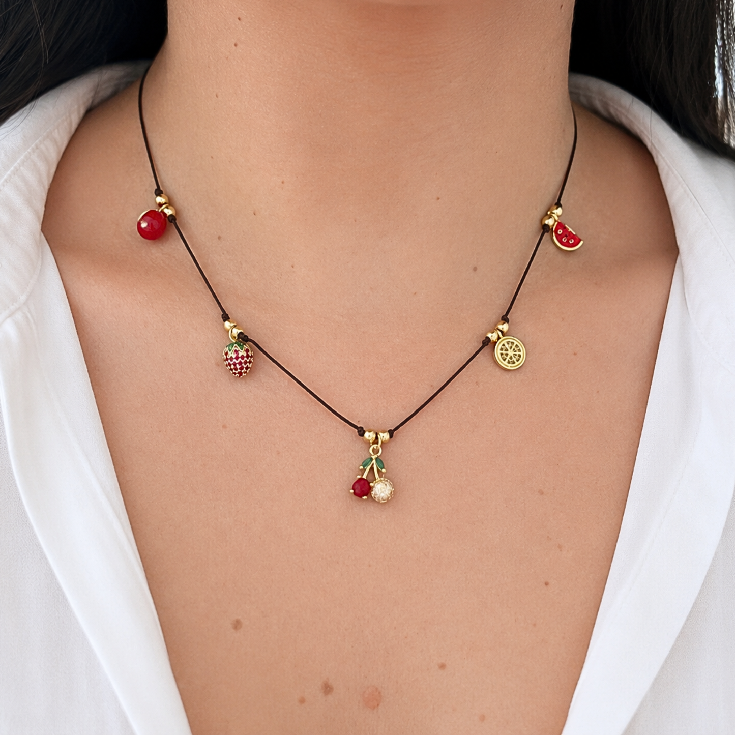 Whimsical fruit charm necklace with a black cord and sparkling gold accents, displayed on a neutral stone background with delicate dried flowers.