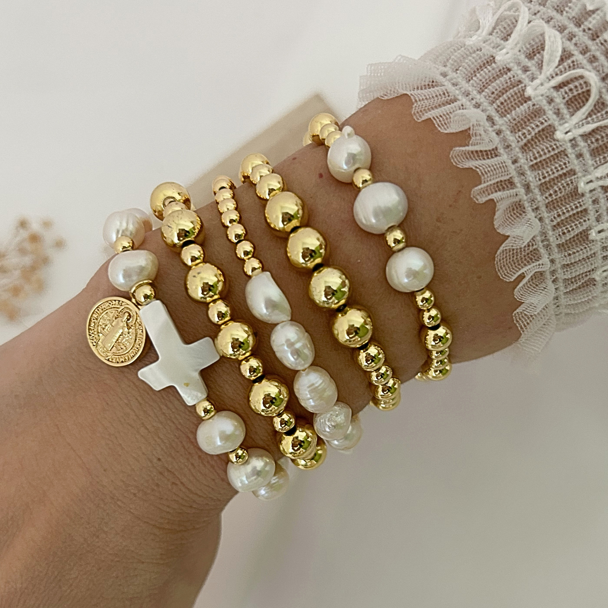 A close-up of a person's wrist wearing five stacked beaded bracelets. The bracelets feature white freshwater pearls, polished gold-tone beads, a white cross charm, and a gold-tone religious medallion.