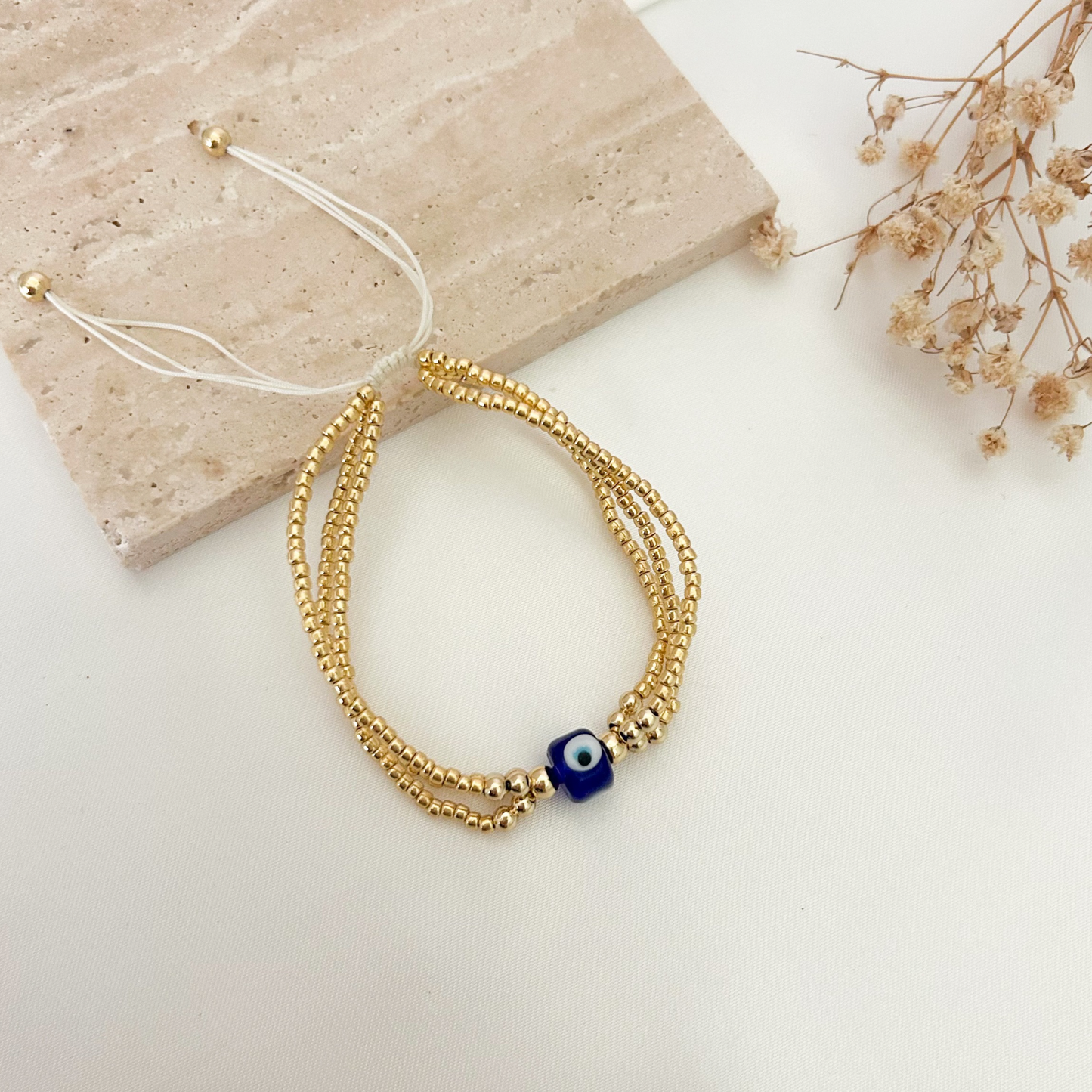 Adjustable handmade bracelet with three strands of gold seed beads and a classic blue evil eye charm, displayed on a neutral background.