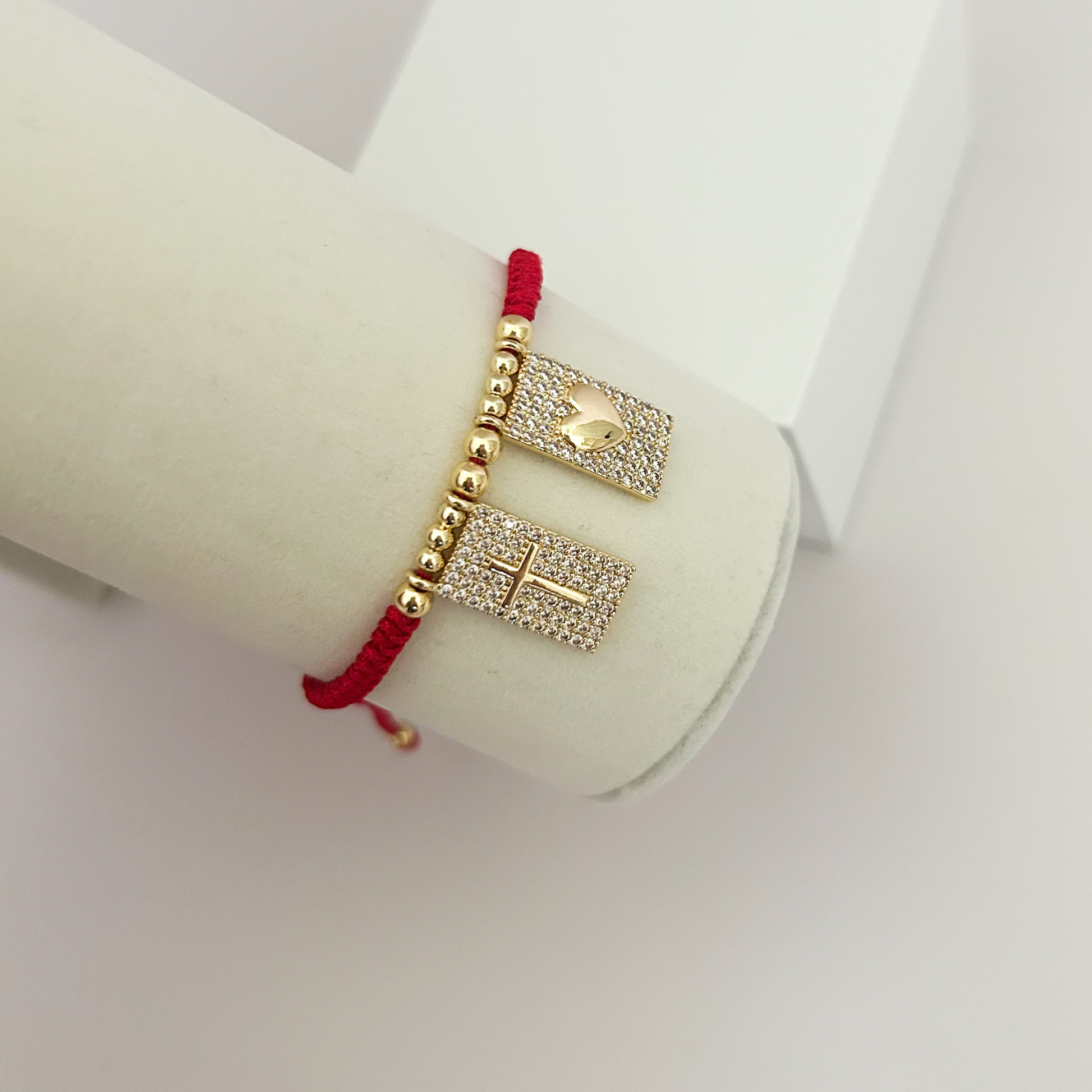 Side view of a red adjustable bracelet with gold-tone cross and heart charms embellished with shimmering crystals, showcased on a jewelry display.