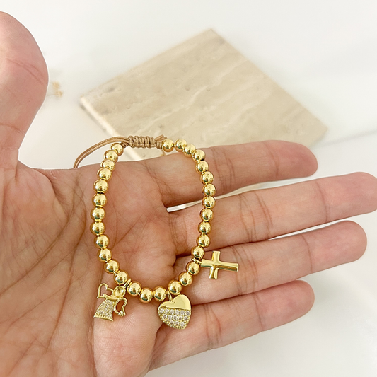 A gold beaded bracelet with a cross, angel, and heart charm, held in a person's hand against a light background.