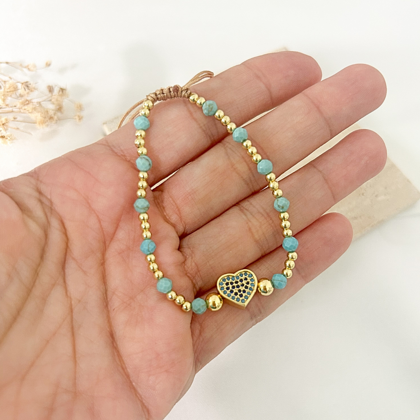 A delicate gold beaded bracelet with light blue turquoise beads and a heart-shaped charm, held in a person's hand.