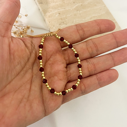 A delicate beaded bracelet with alternating gold-toned beads and deep red faceted beads, held in a person's hand.