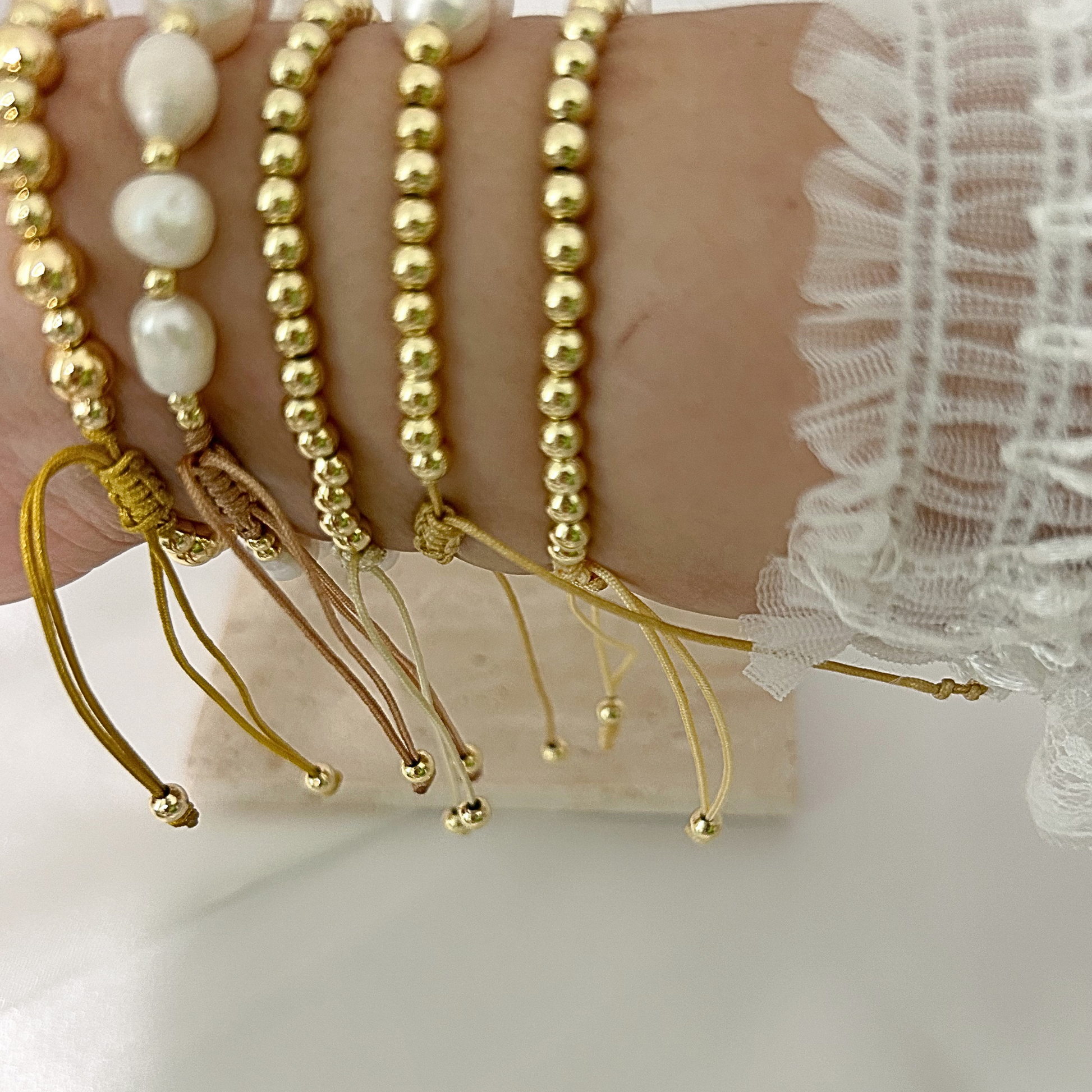A close-up of a person's wrist wearing multiple gold-toned beaded bracelets, including one with large white pearl-like beads and two with thin braided cords and small gold beads at the ends.