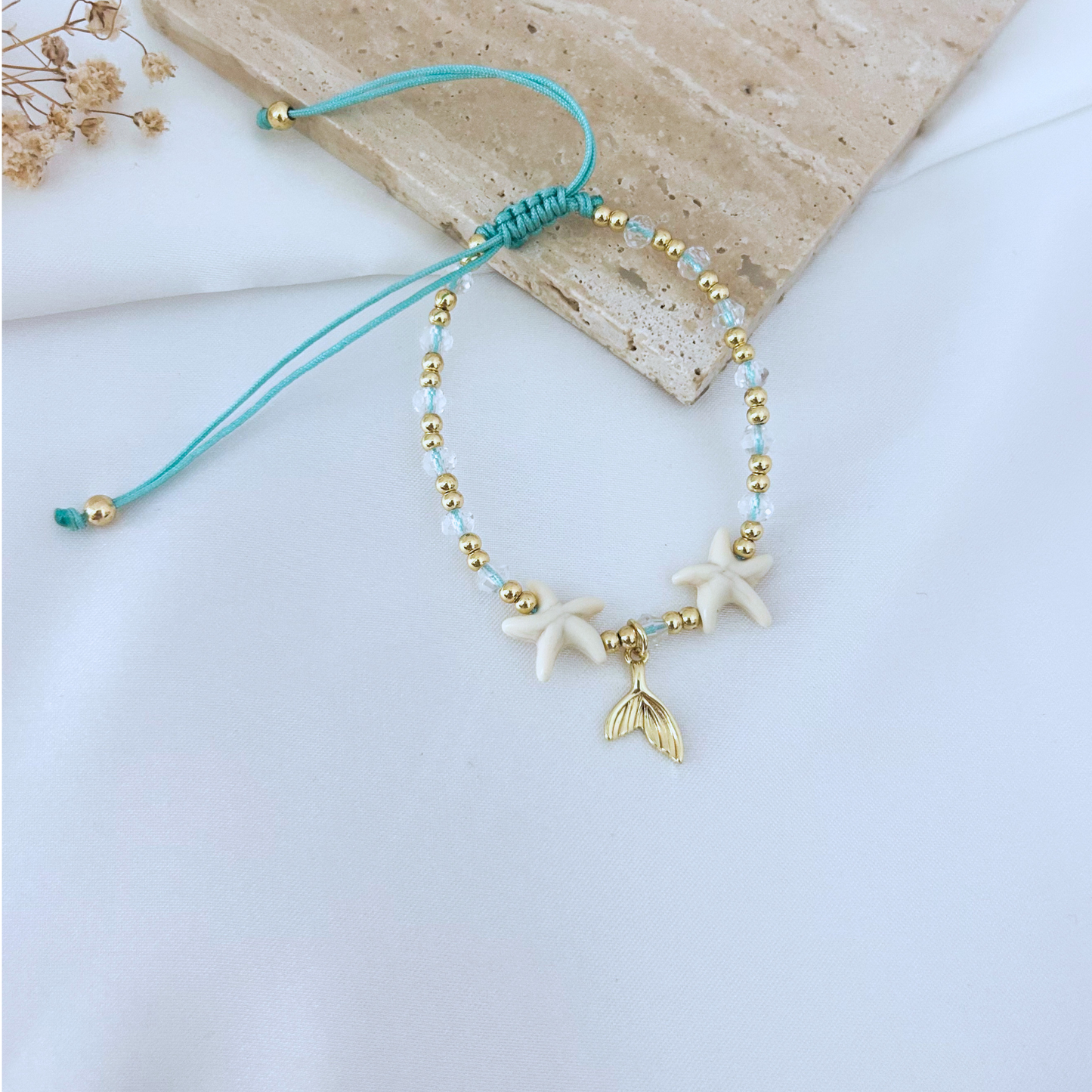 Beach-themed adjustable bracelet displayed on a neutral stone surface, combining white starfish beads, sparkling crystal details, gold beads, and a gold mermaid tail charm. A playful and elegant accessory inspired by the ocean.