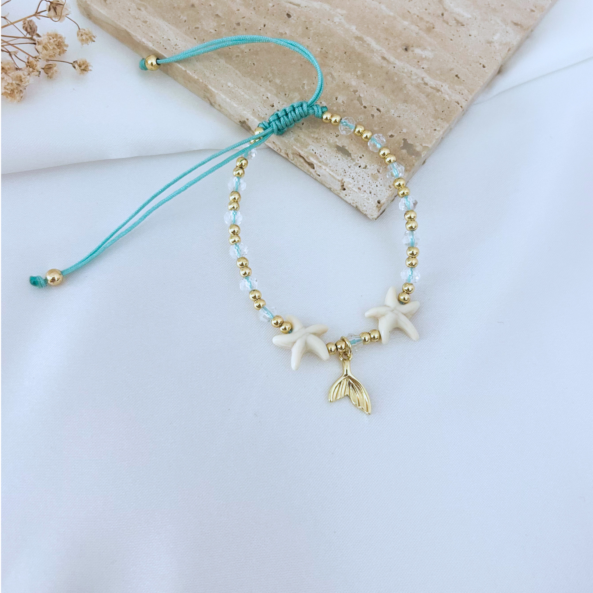Beach-themed adjustable bracelet displayed on a neutral stone surface, combining white starfish beads, sparkling crystal details, gold beads, and a gold mermaid tail charm. A playful and elegant accessory inspired by the ocean.