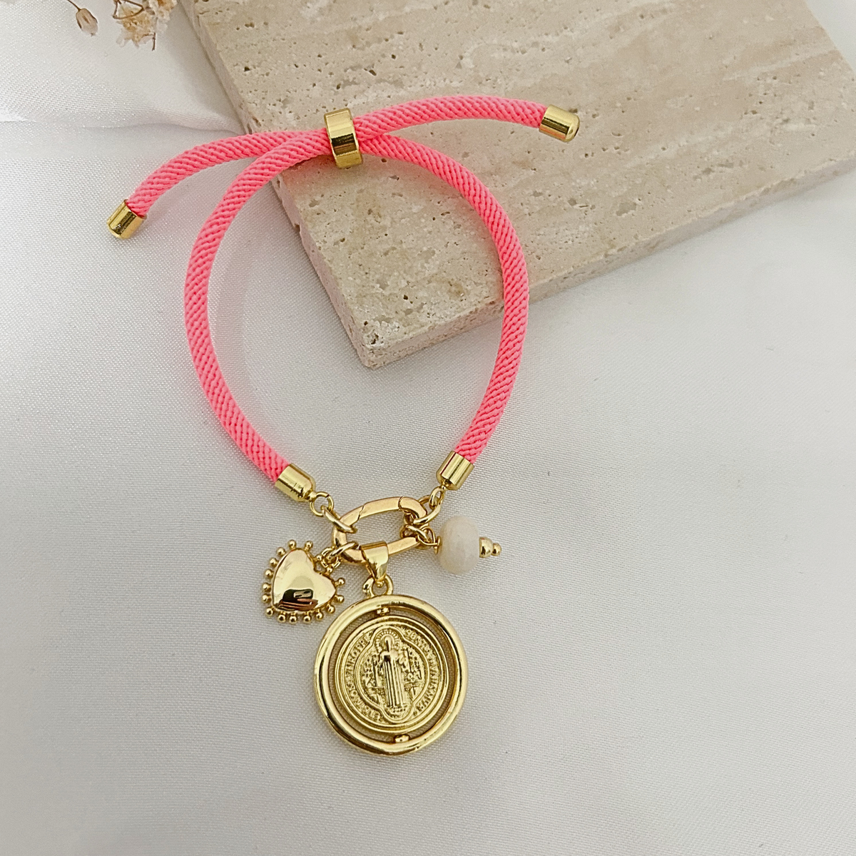 Neon pink charm bracelet with spinning Saint Benedict medal, heart, and white bead on a stone tray