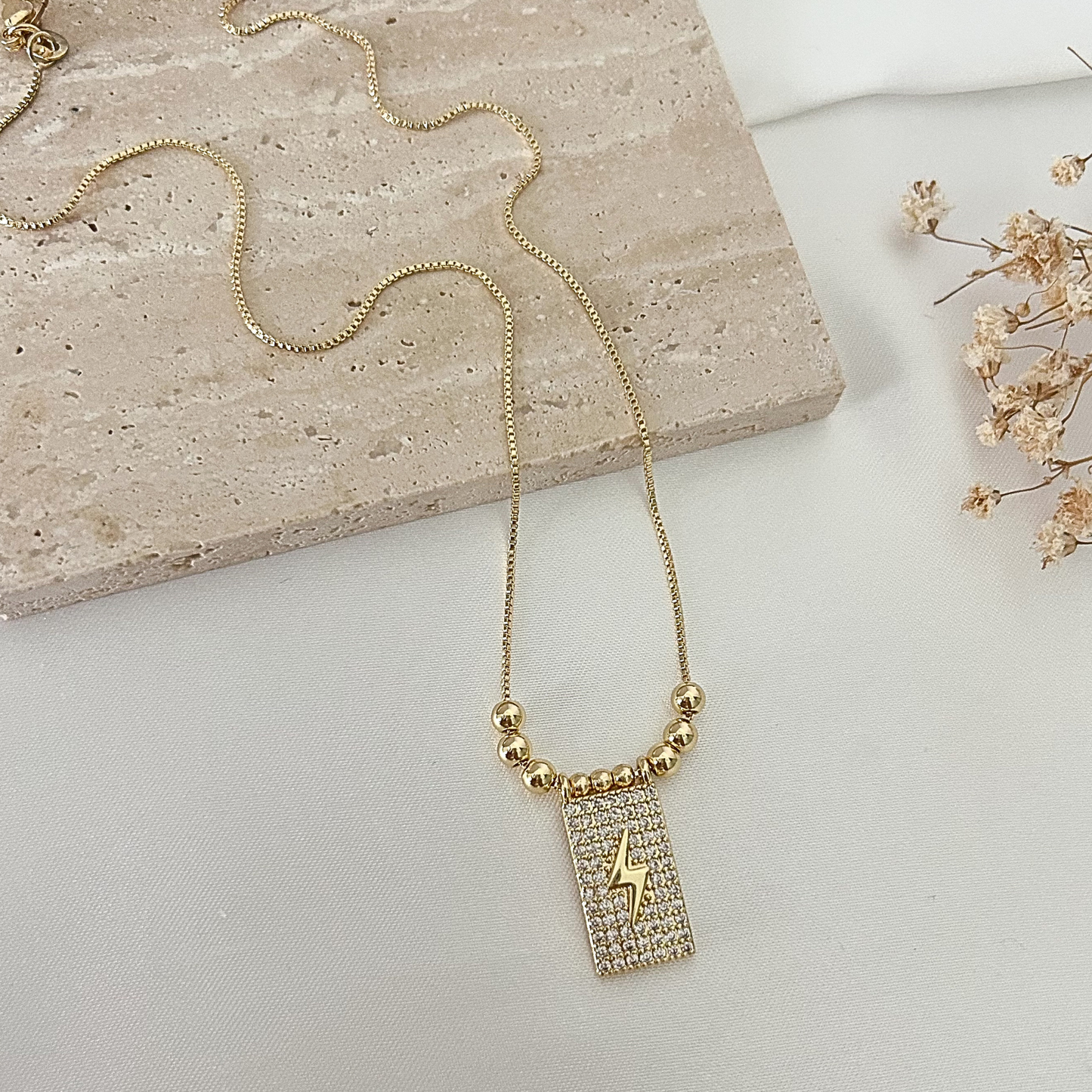 Gold-tone adjustable necklace featuring a sparkling rectangular pendant with a lightning bolt design, accented with polished gold beads and displayed on a neutral stone surface.