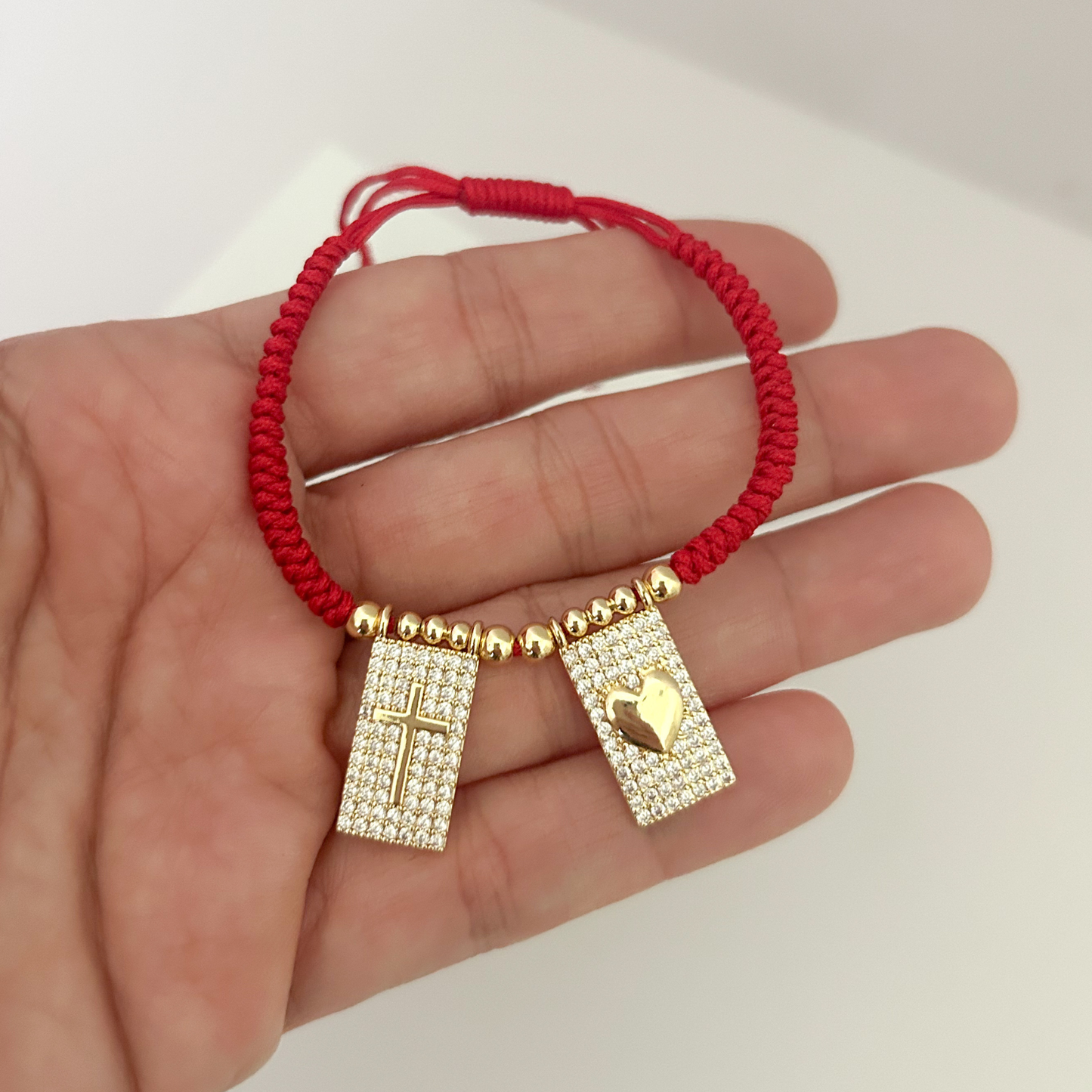 Handheld view of an adjustable red bracelet with sparkling gold-tone cross and heart charms, emphasizing its size, craftsmanship, and everyday wearability.