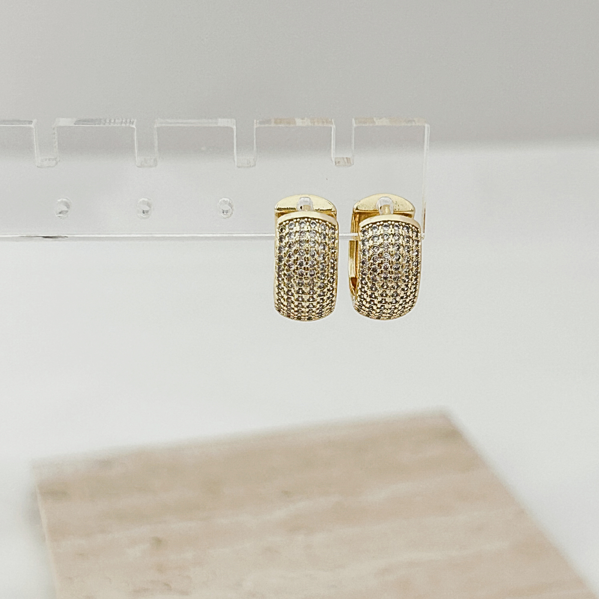 A pair of gold hoop earrings covered in small pavé-set clear stones, displayed on a clear acrylic stand against a light gray background.