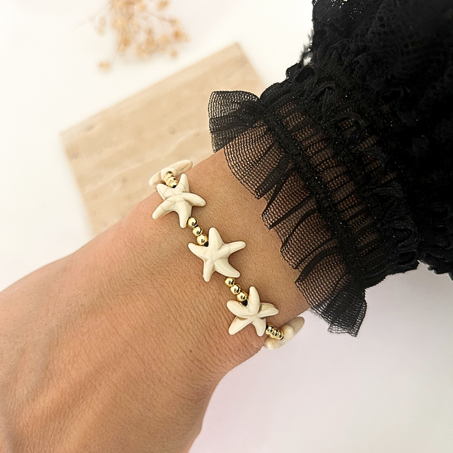 A close-up of a person's wrist wearing a delicate bracelet with three cream-colored starfish charms and small gold beads, paired with a black ruffled sleeve.