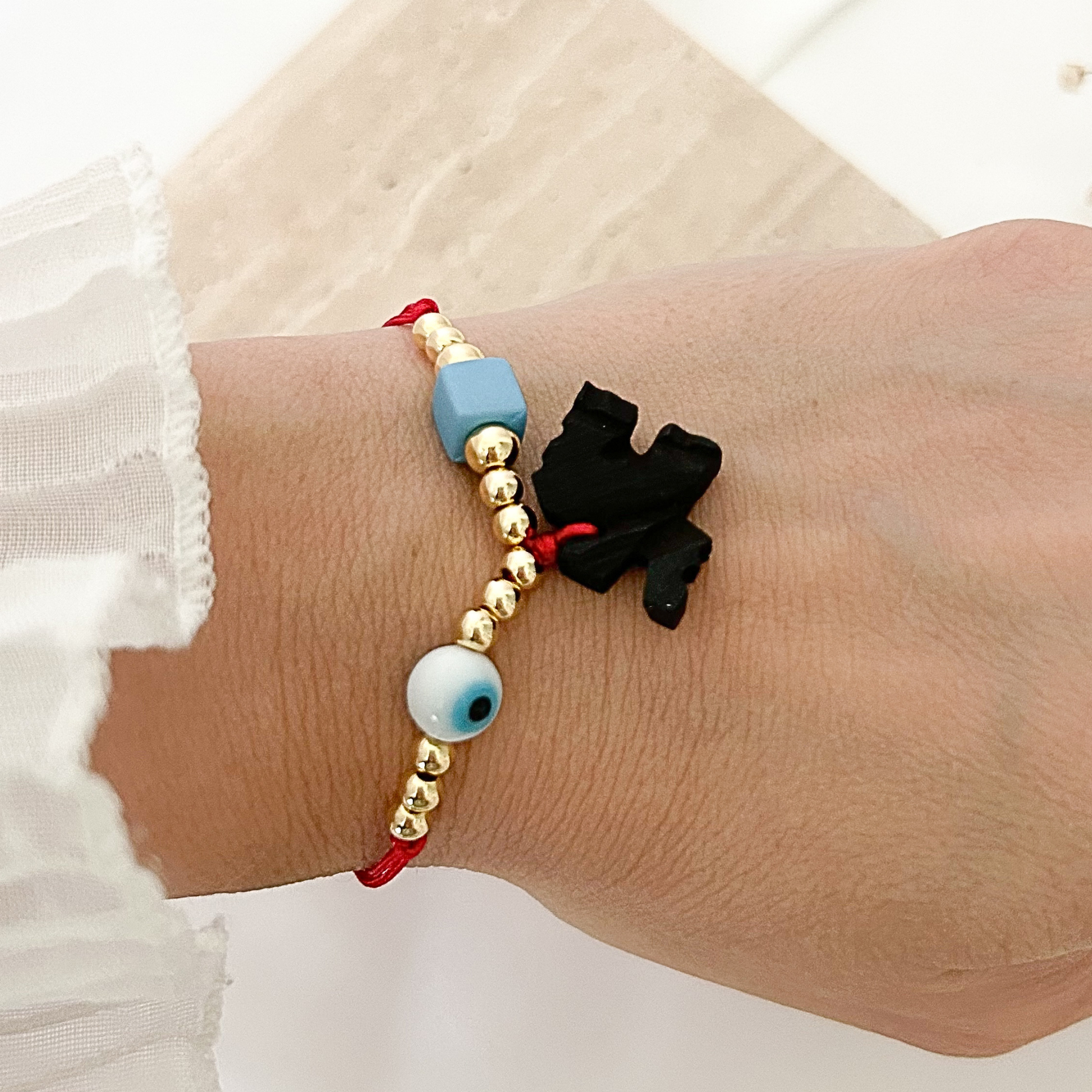 Red adjustable bracelet featuring gold-tone beads, a blue evil eye bead, a geometric blue accent bead, and a carved black elephant charm, styled on the wrist with a soft white sleeve.