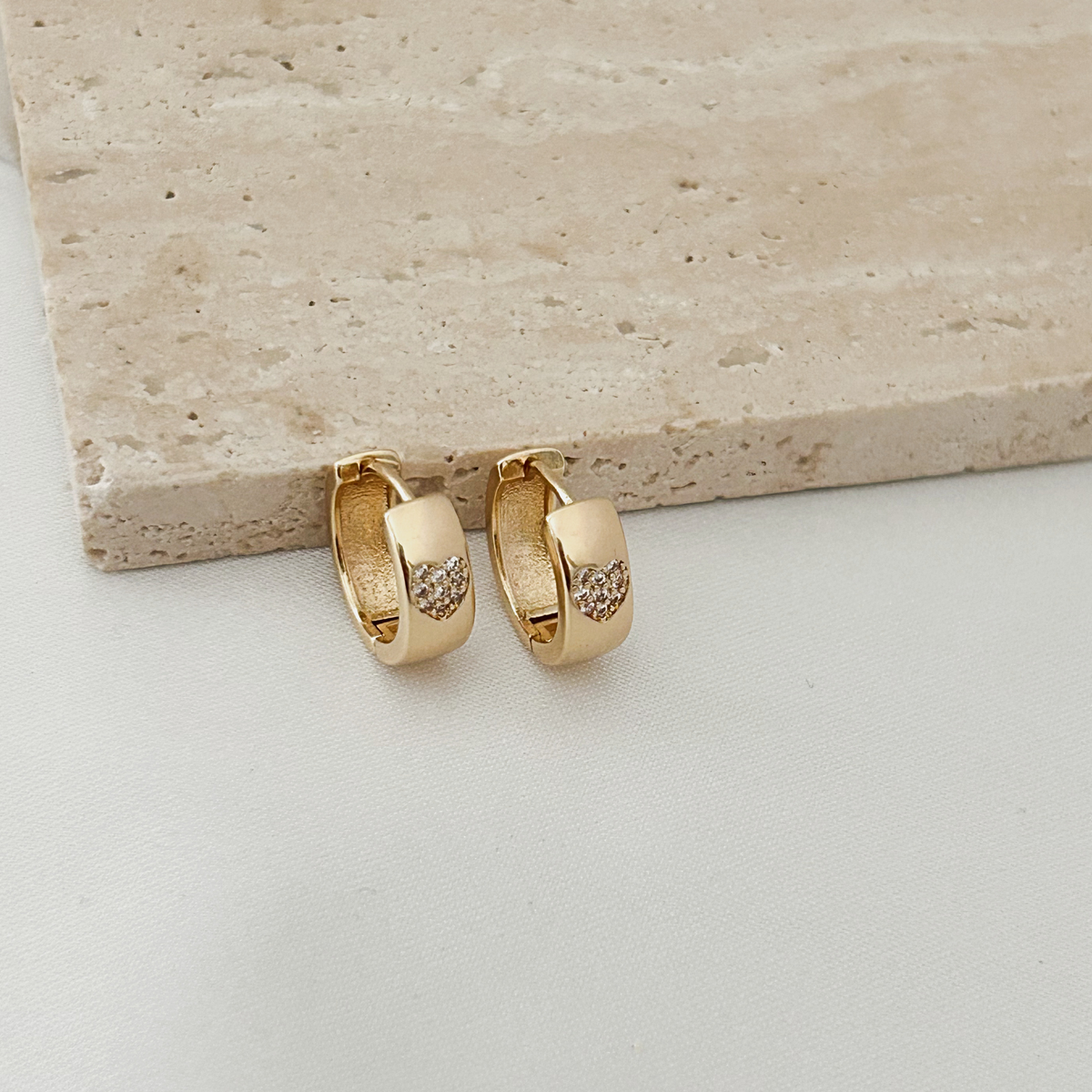 A pair of small gold huggie hoop earrings with a textured inlay and small clear stones, displayed on a light beige stone slab over a white surface.