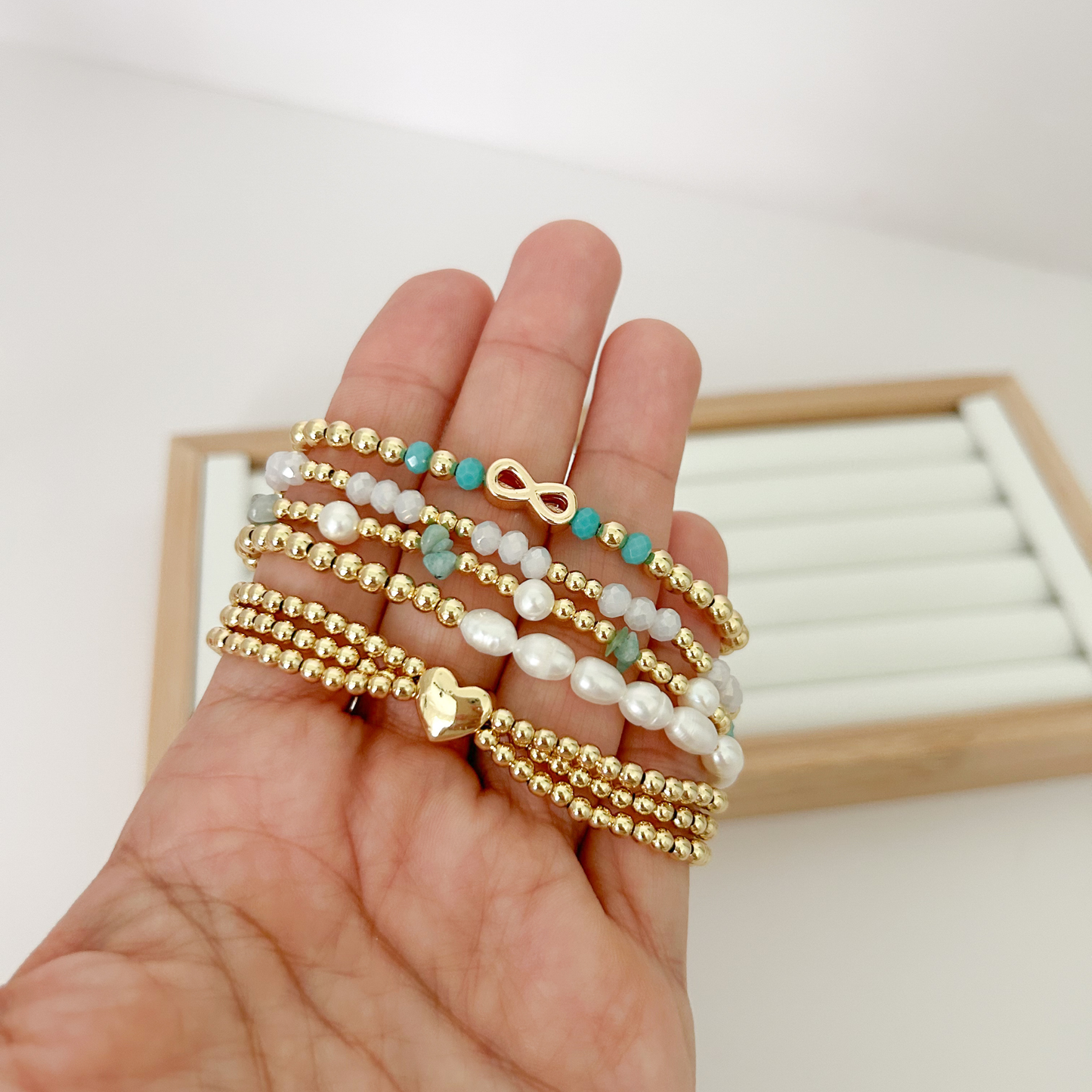 Five gold beaded bracelets with turquoise stones and infinity charm held in hand