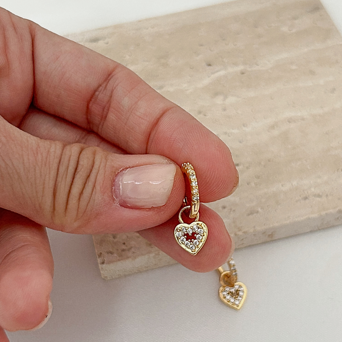 A close-up of a hand holding a small gold heart charm with a red heart in the center and clear stones around it, with another matching charm hanging below, against a light stone slab background.