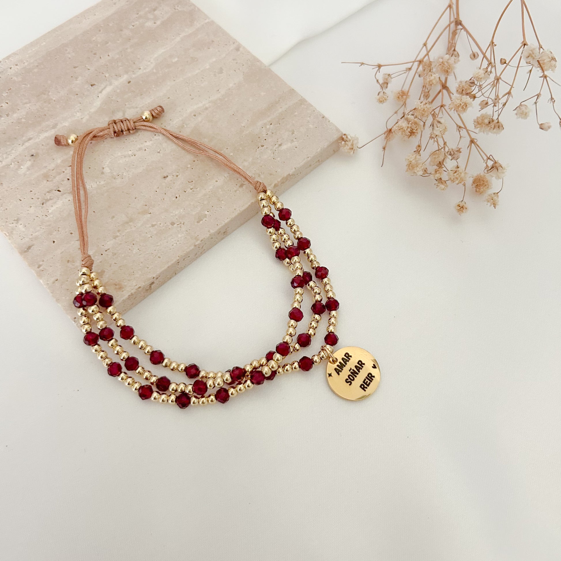 Adjustable handmade bracelet with three rows of burgundy faceted beads, gold-tone seed beads, and an inspirational engraved charm reading “Amar, Soñar y Reír,” displayed on a neutral backdrop.