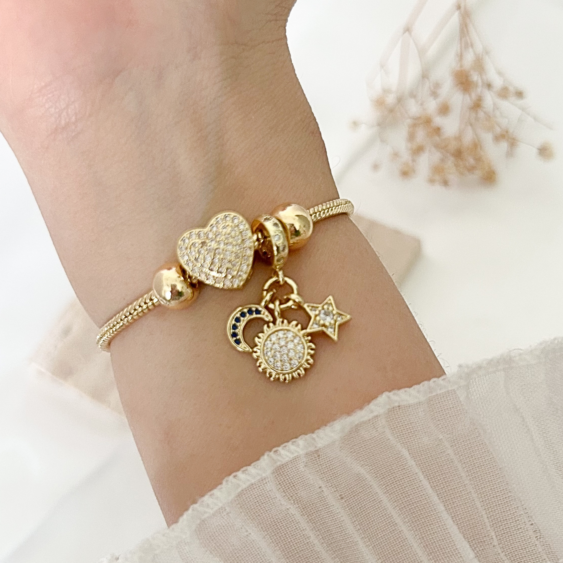Elegant gold bracelet worn on the wrist, showcasing a pavé heart charm alongside detailed sun, moon, and star pendants, creating a celestial-inspired jewelry design.