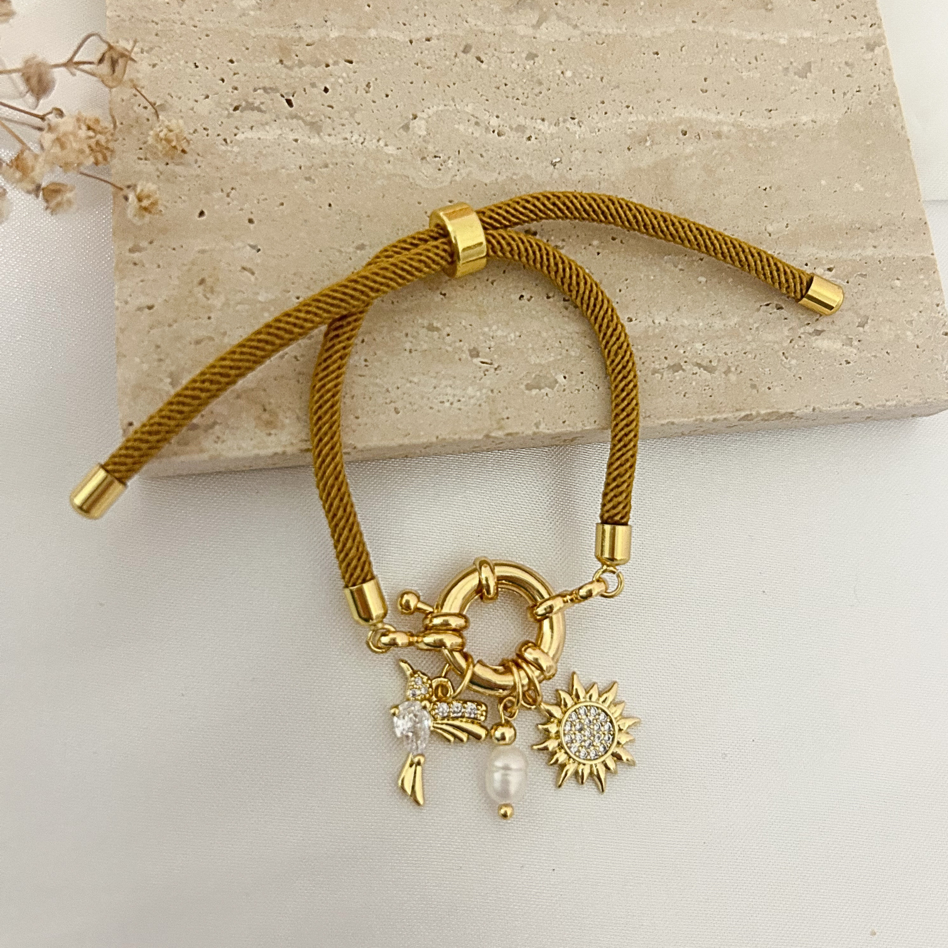 A gold-toned braided cord bracelet with a toggle clasp and three dangling charms: a sun, a crescent moon with a flower, and a pearl bead, displayed on a light stone slab.