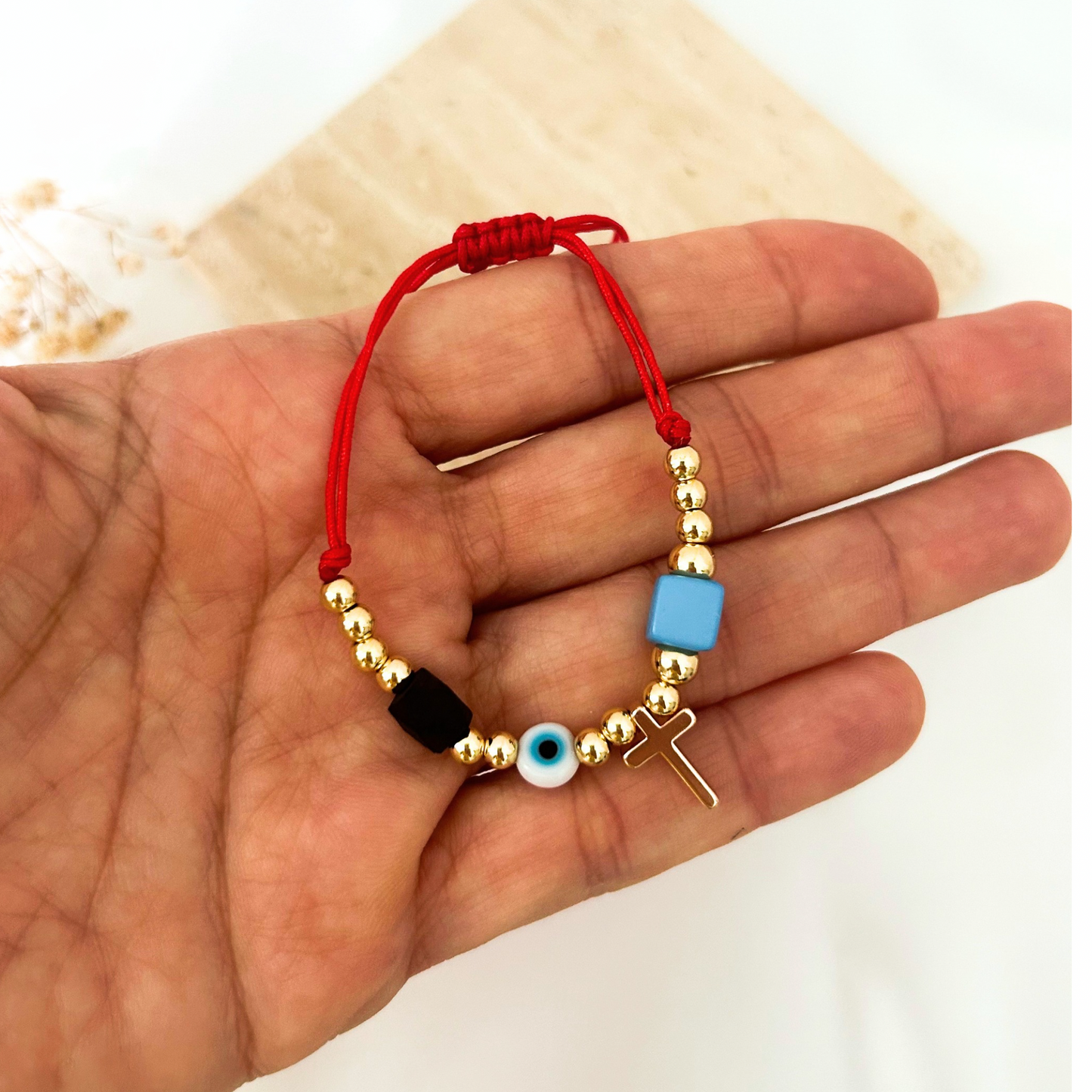 Close-up of an adjustable red string bracelet with polished gold beads, a dangling gold cross, a classic blue evil eye bead, and geometric black and blue accent beads. Lightweight and perfect for daily wear.