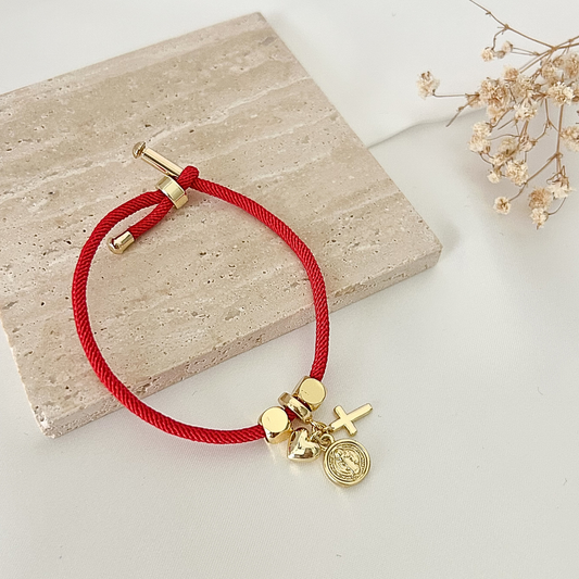 Faith-inspired red bracelet with gold accents, including a cross charm, heart charm, and Saint Benedict medallion, displayed on a stone surface.