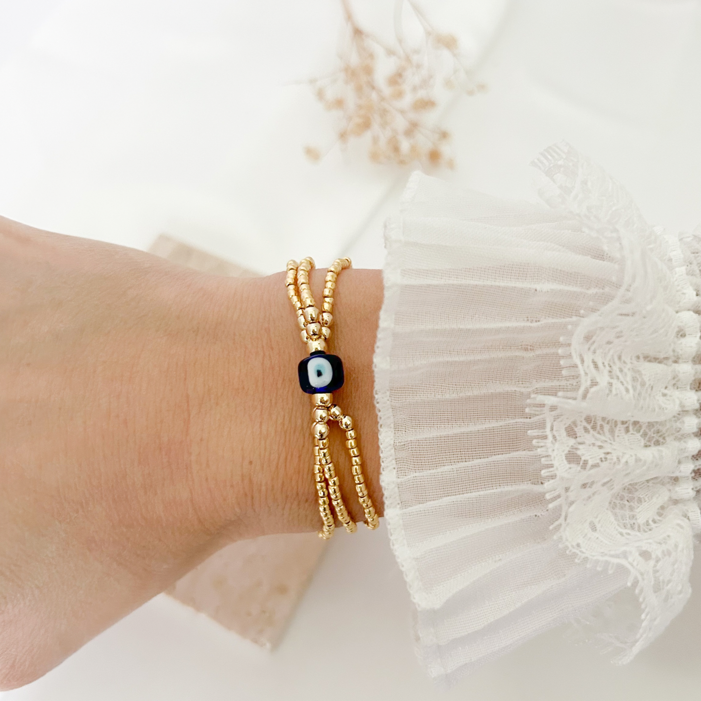 Delicate triple-strand gold beaded bracelet featuring a blue evil eye accent bead, worn on the wrist with a feminine lace sleeve.
