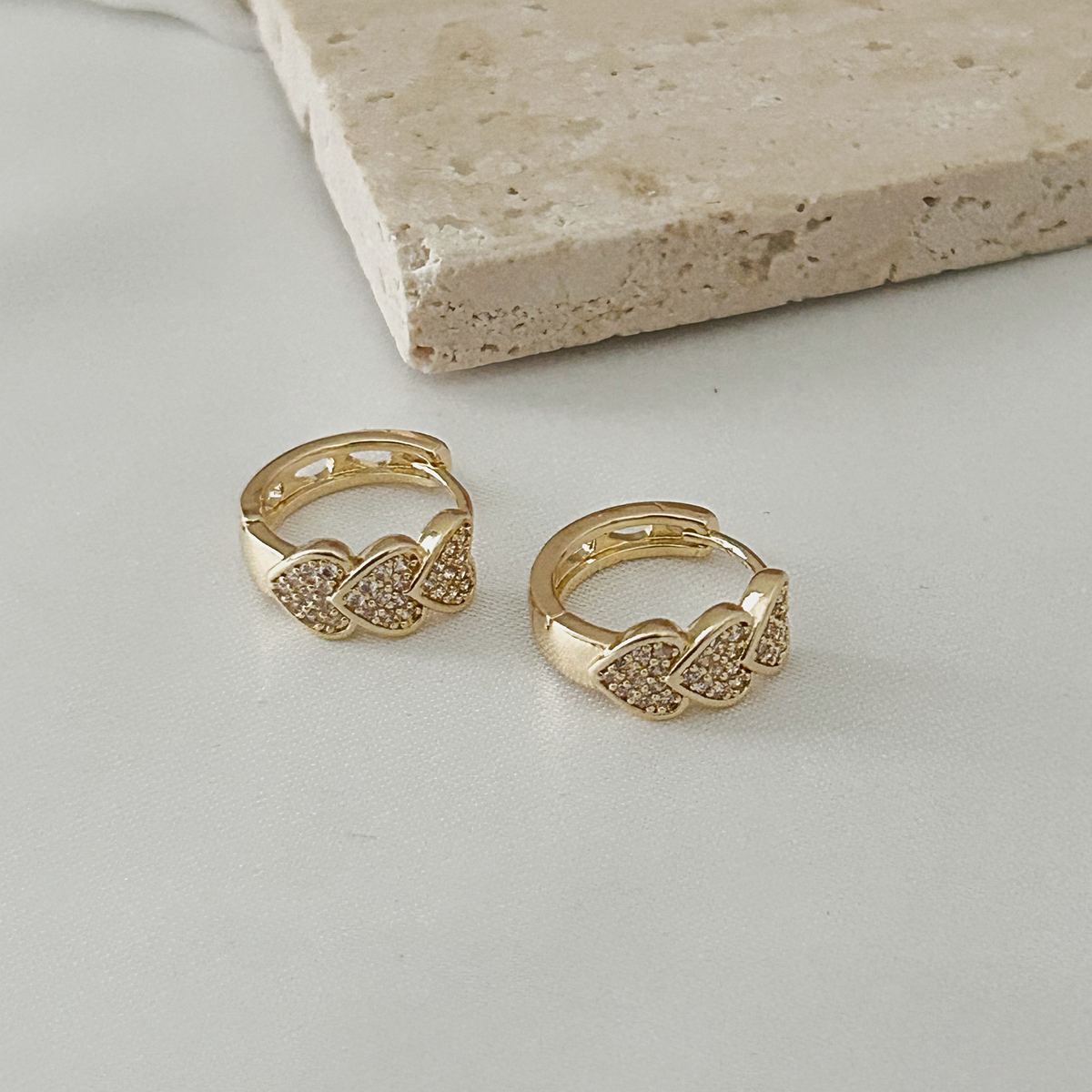 A pair of gold huggie hoop earrings with pavé-set clear stones in a wing-like design, displayed on a light gray surface with a beige stone prop in the background.