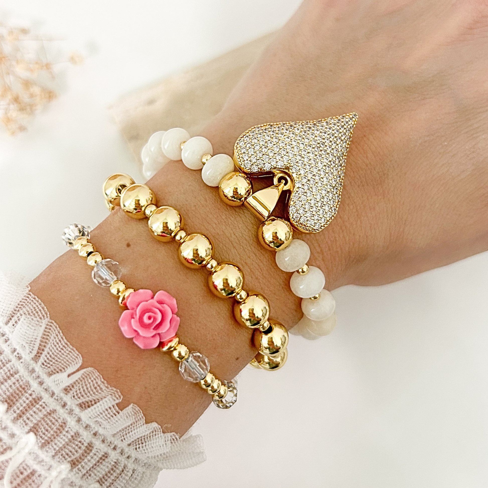 A close-up of a wrist wearing three stacked bracelets: a gold beaded bracelet, a white beaded bracelet with a rhinestone heart charm, and a gold beaded bracelet with a pink rose charm.