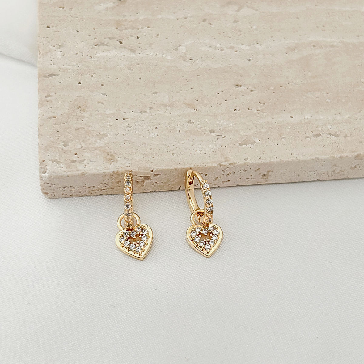 A pair of gold hoop earrings with dangling heart charms paved with small clear stones, displayed on a light beige stone slab over a white surface.