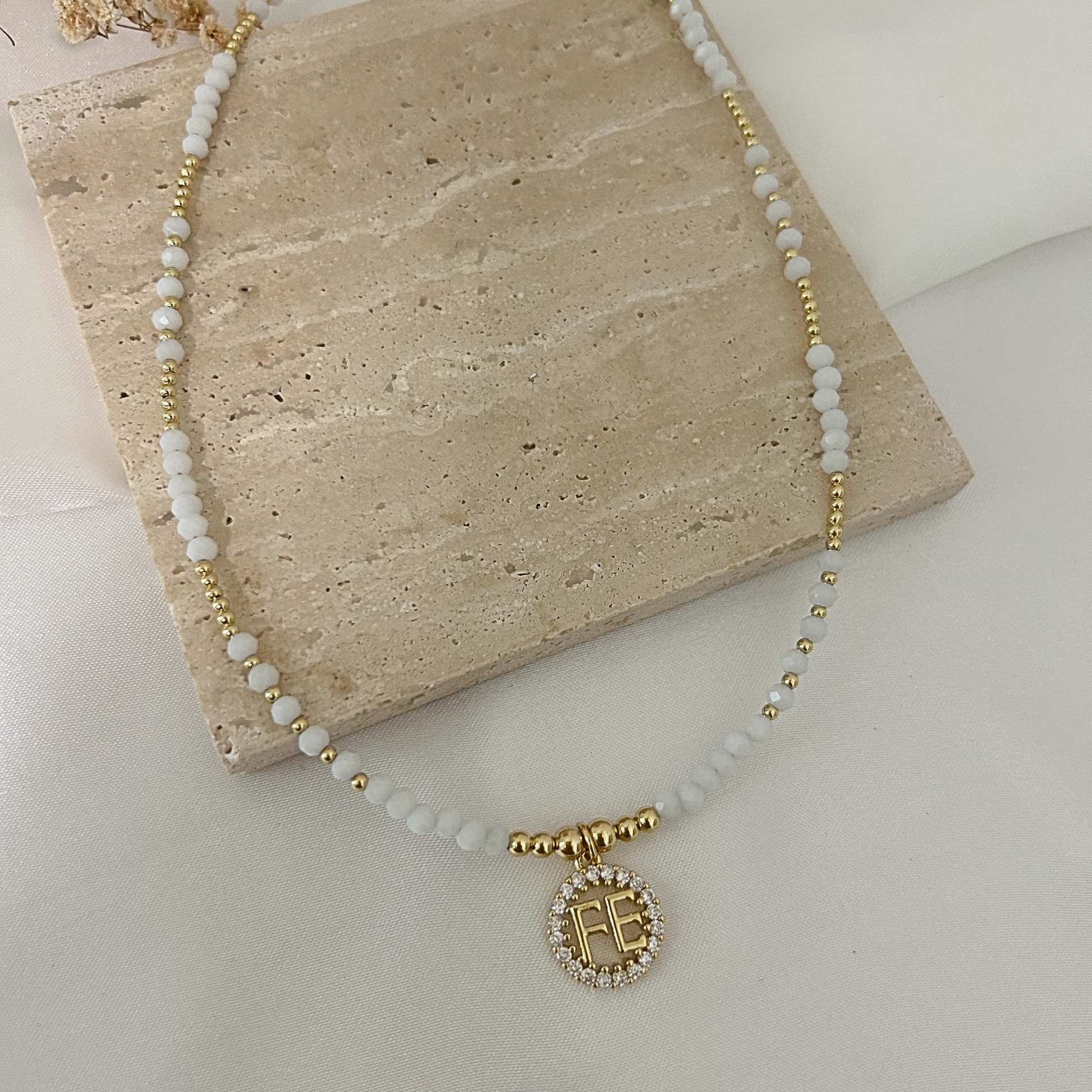 Collar de cuentas blancas y doradas con medalla redonda que dice “FE”, rodeada de circonias brillantes, presentado sobre un fondo neutro de estilo minimalista.