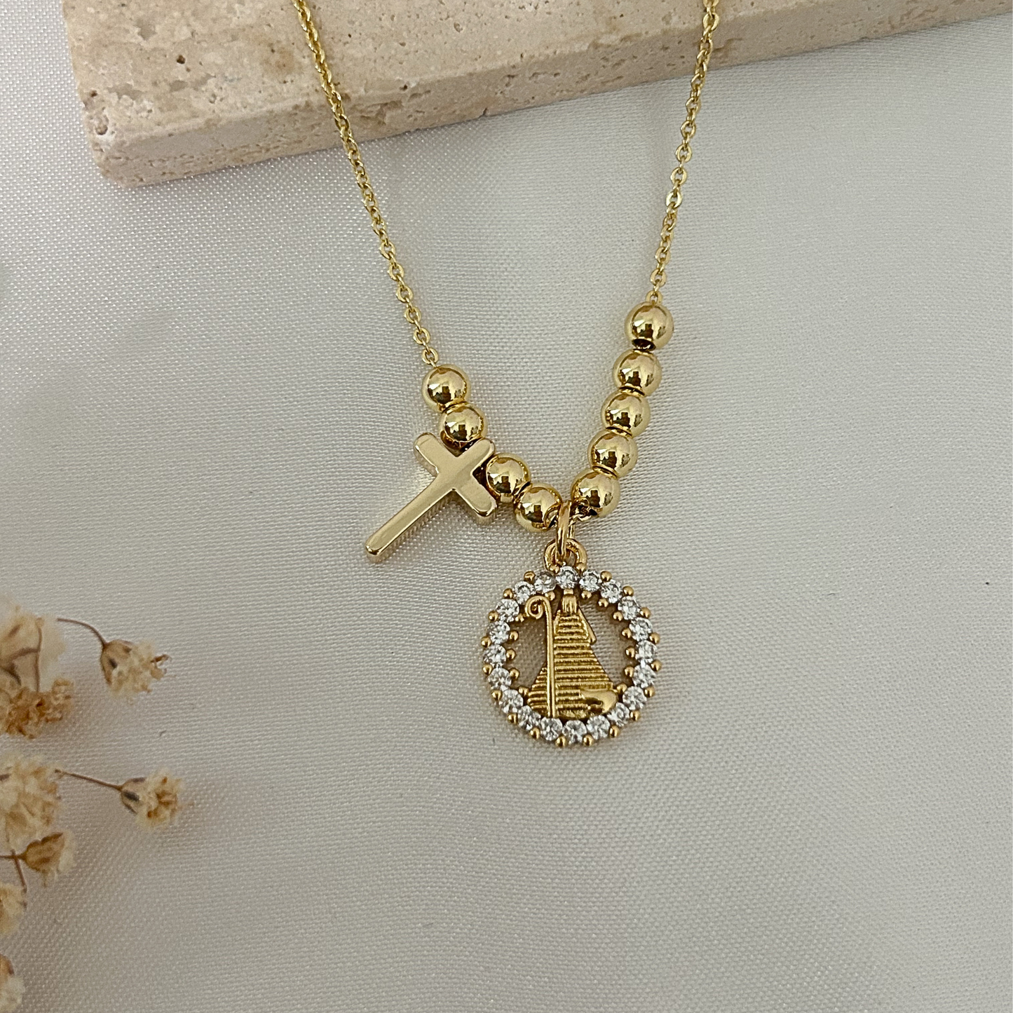 Primer plano de un collar dorado con cruz y medalla de la Divina pastora con halo de circonias, fotografiado sobre una superficie de tela clara con una base de piedra beige.