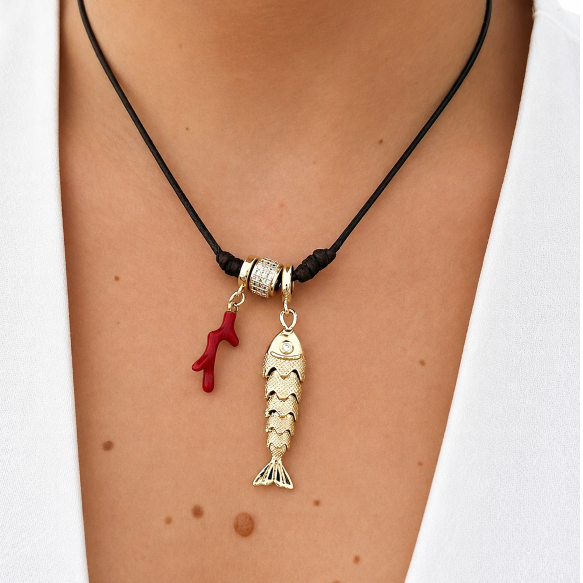 Model wearing an adjustable black cord necklace with a dangling gold fish charm, red coral accent, and gold crystal bead. A chic ocean-inspired statement piece that adds a touch of coastal elegance to any outfit.